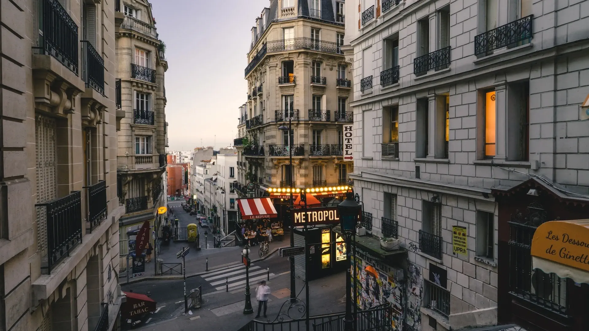 Vue panoramique d'un quartier pittoresque de Paris avec des bâtiments historiques aux façades colorées, des rues pavées animées de passants et de petits cafés, et la Tour Eiffel en arrière-plan sous un ciel bleu clair.